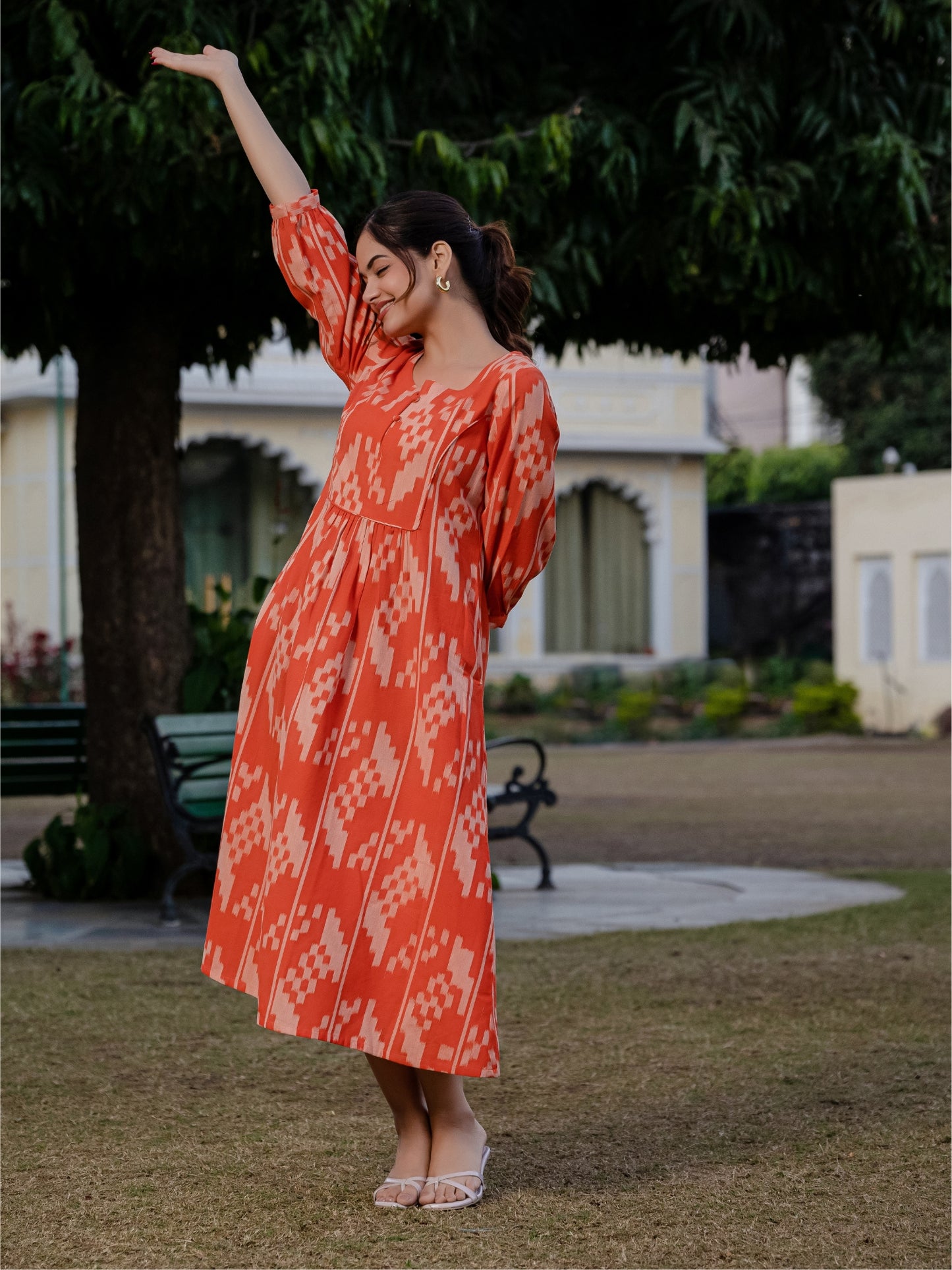 A woman in a vibrant orange ikkat printed dress turns slightly, offering a side-back view in a serene garden setting.