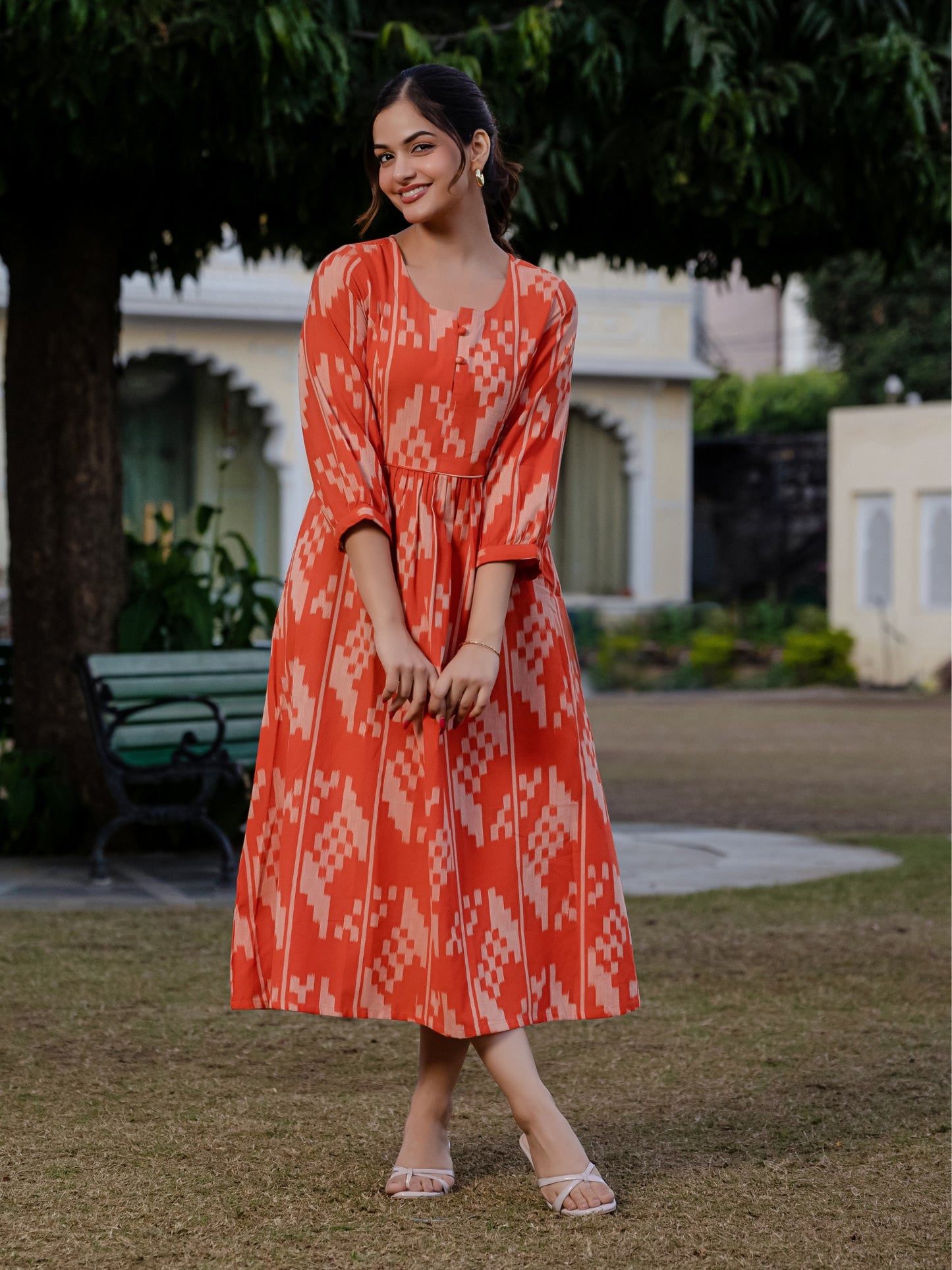 A woman wearing an orange ikkat cotton dress poses outdoors in a garden setting, smiling softly with hands clasped in front.
