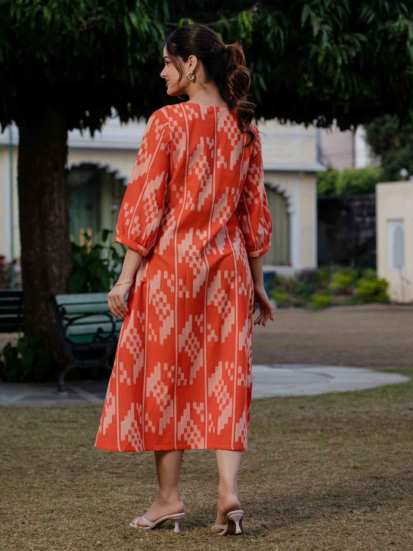 Back view of an orange ikkat midi dress worn by a woman standing outdoors, highlighting the fabric pattern and dress silhouette.