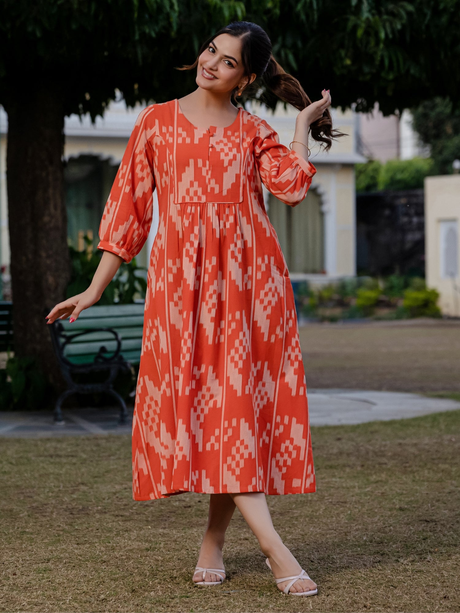 Full-length outdoor photo of a woman in an orange ikkat printed dress, standing on grass with trees and heritage-style architecture in the background.